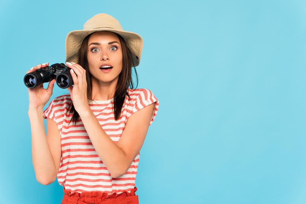 young surprised woman in hat holding binoculars isolated on blue - Photo, Image
