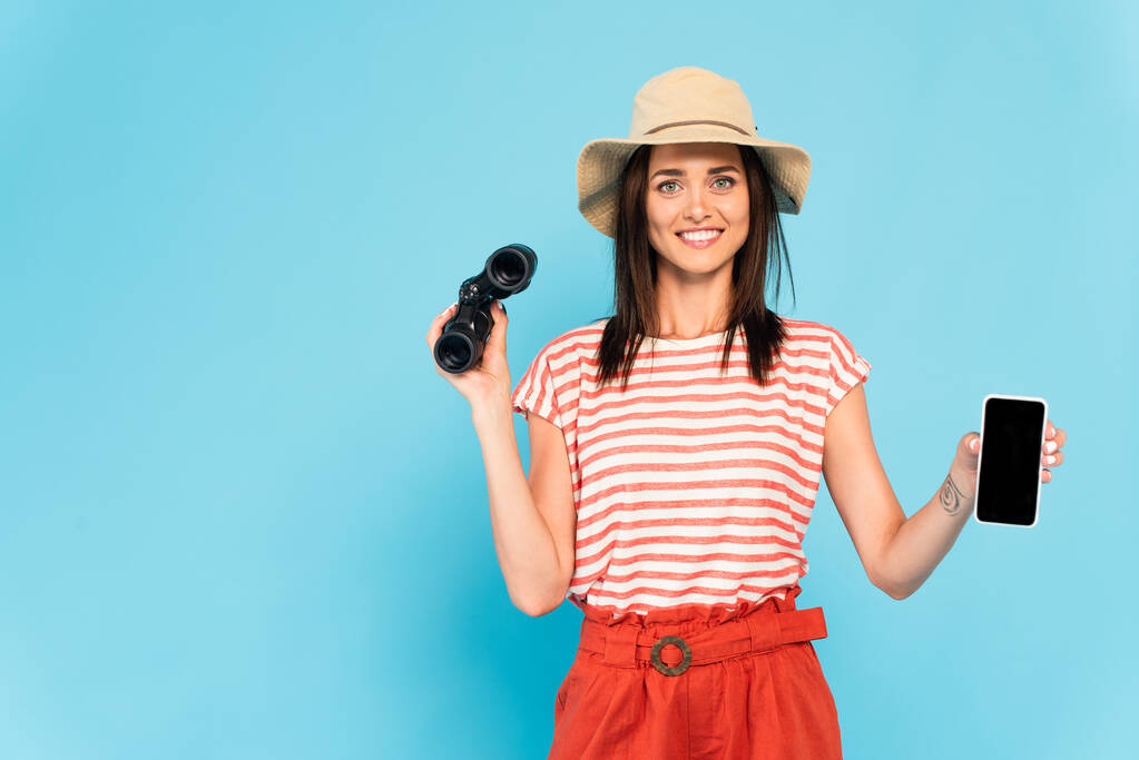 happy woman in hat holding smartphone with blank screen and binoculars on blue - Photo, Image