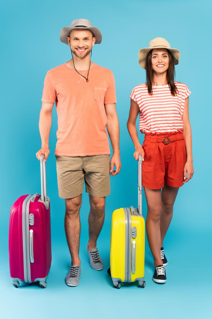 feliz pareja en sombreros de pie cerca de bolsas de viaje en azul  - Foto, imagen