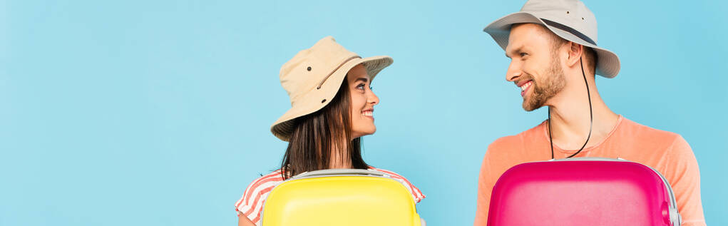 панорамный снимок счастливой пары, смотрящей друг на друга и держащей багаж, изолированный на голубом  - Фото, изображение