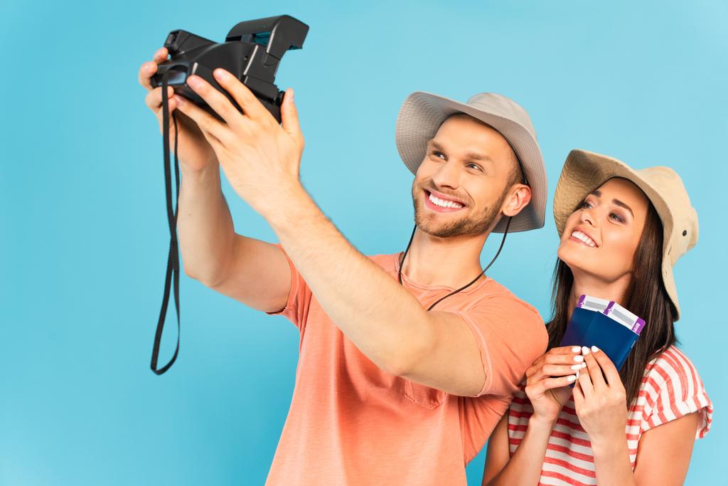 Şapkalı mutlu bir adam klasik fotoğraf makinesi tutuyor ve mavi üzerine izole edilmiş neşeli bir kızla selfie çekiyor. - Fotoğraf, Görsel