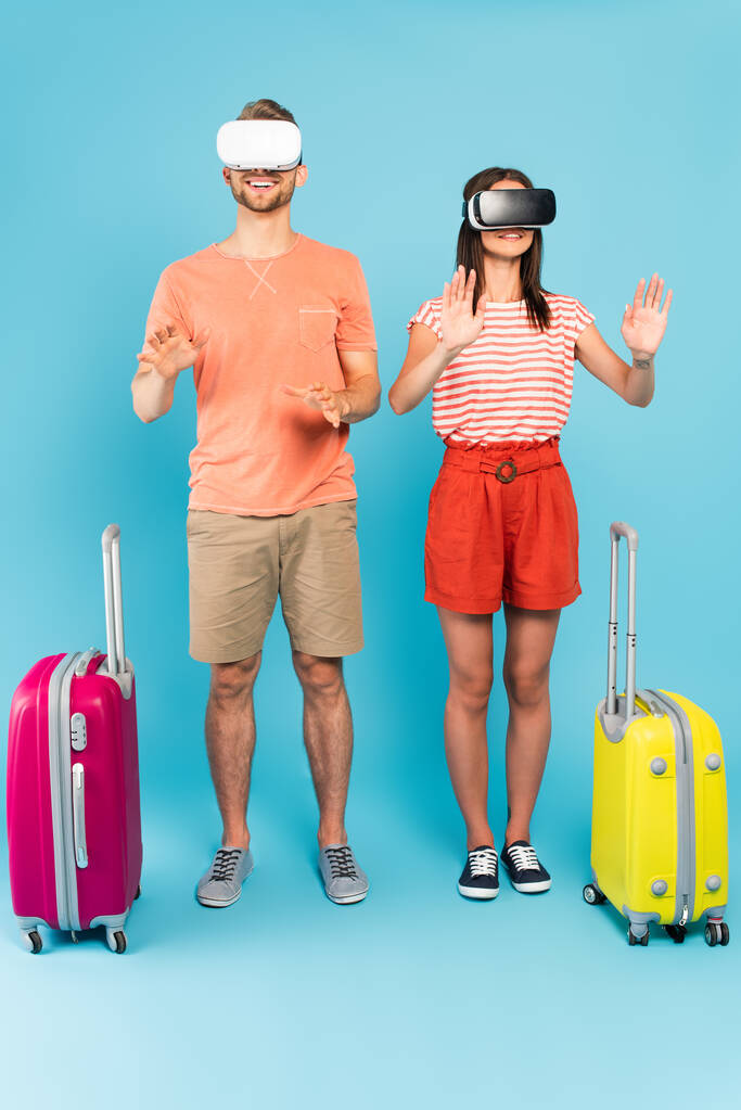 wesoła para w wirtualnej rzeczywistości zestawy słuchawkowe stojące z bagażem na niebiesko  - Zdjęcie, obraz