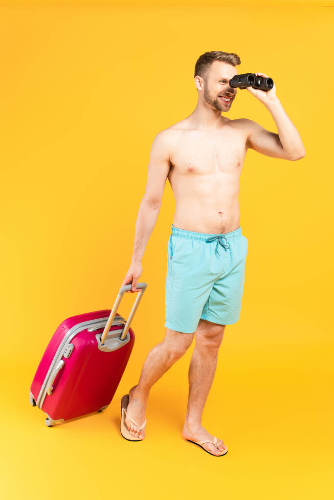 ευτυχισμένος άνθρωπος κοιτάζοντας μέσα από κιάλια και στέκεται με αποσκευές στο κίτρινο - Φωτογραφία, εικόνα