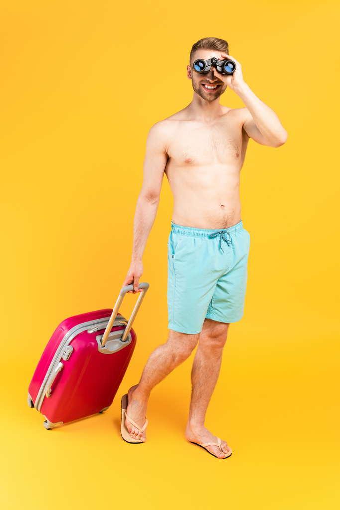 щасливий м'язистий чоловік дивиться через бінокль і стоїть з багажем на жовтому
 - Фото, зображення