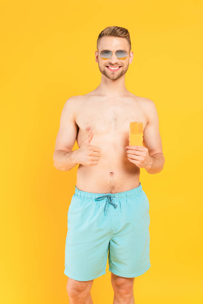 gelukkig shirtloze man in zonnebril tonen duim omhoog en houden fles met zonnebrand geïsoleerd op geel  - Foto, afbeelding