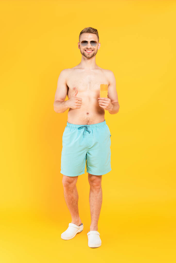 gelukkig shirtloze man in zonnebril tonen duim omhoog en houden fles met zonnebrandcrème op geel  - Foto, afbeelding