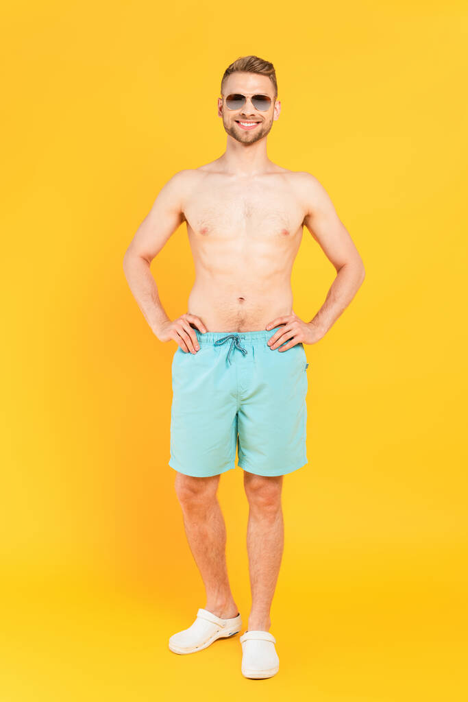 hombre feliz sin camisa en gafas de sol de pie con las manos en las caderas en amarillo  - Foto, imagen