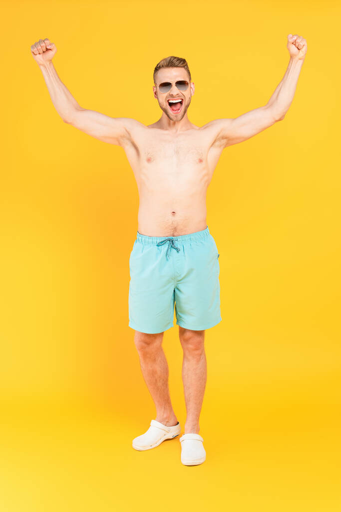 podniecony i bez koszulki mężczyzna w okularach przeciwsłonecznych stojący z dłońmi nad głową na żółtym  - Zdjęcie, obraz