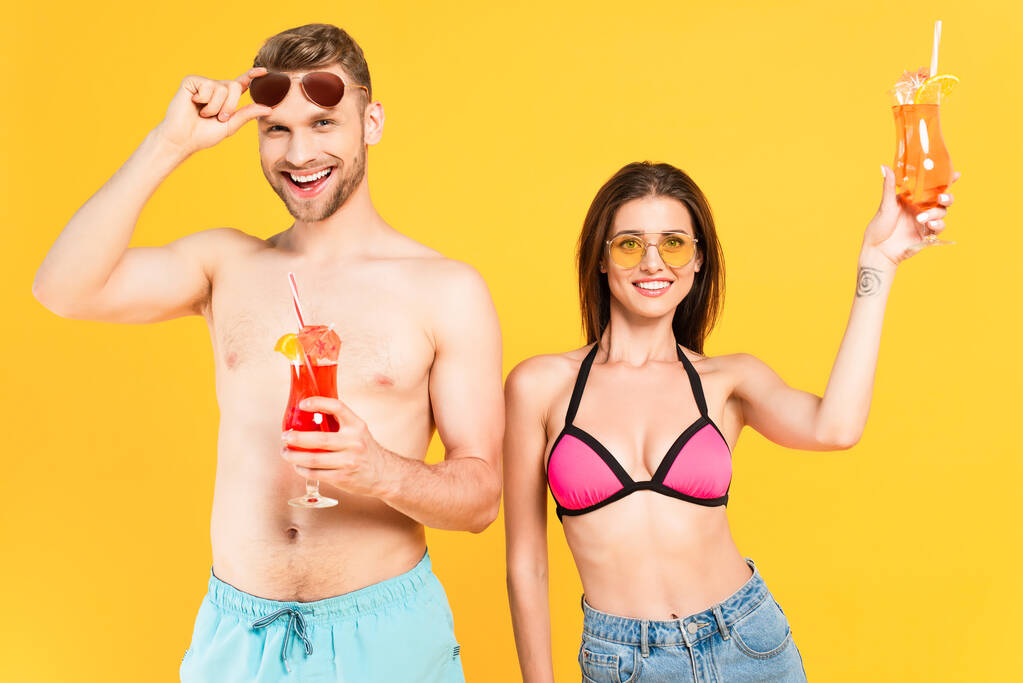 alegre pareja en gafas de sol y trajes de baño celebración cócteles aislados en amarillo - Foto, imagen