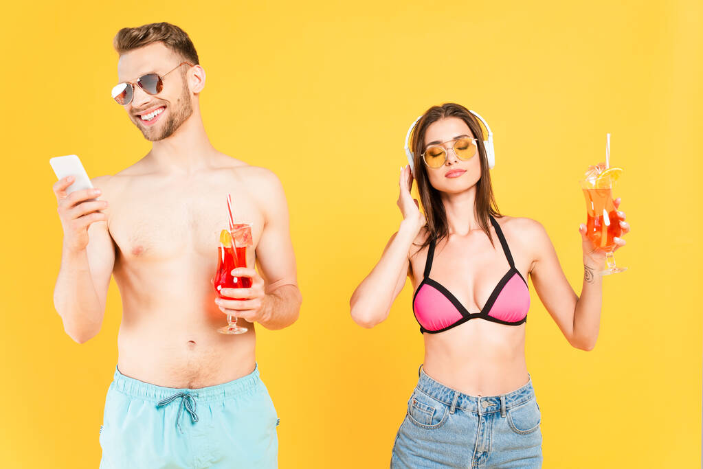 happy man in sunglasses using smartphone and holding cocktail while girl in swimwear listening music isolated on yellow - Photo, Image