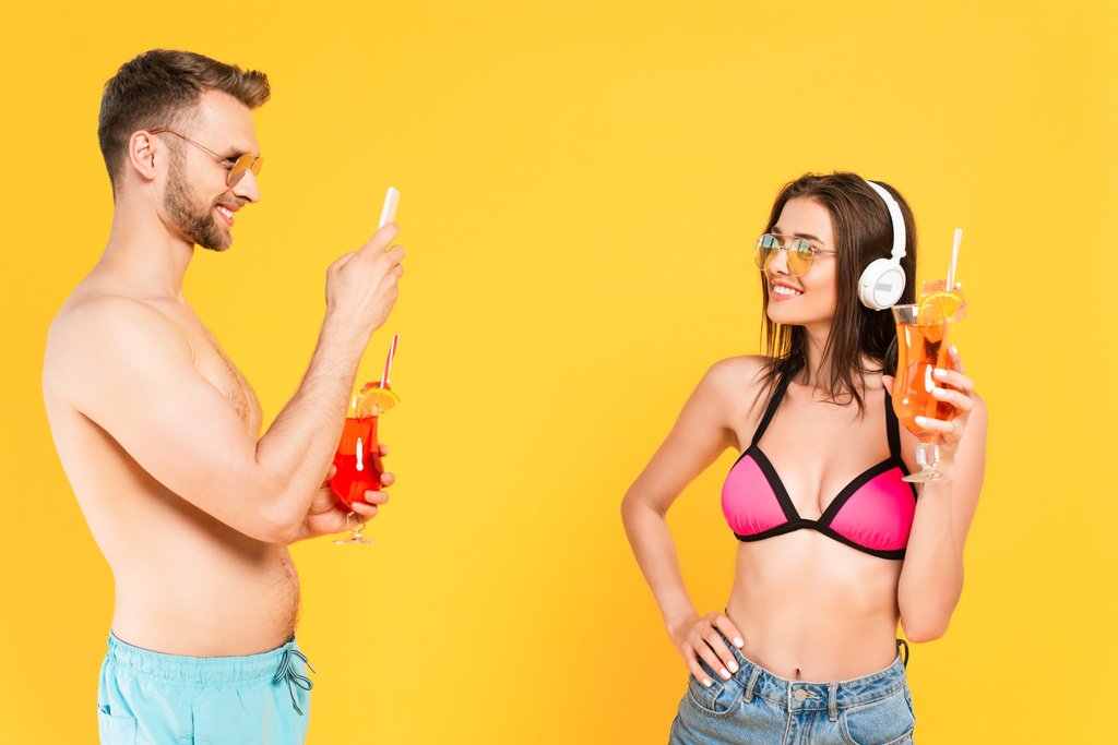 vousatý muž fotografování šťastné dívky ve sluchátkách s koktejlem izolované na žluté - Fotografie, Obrázek