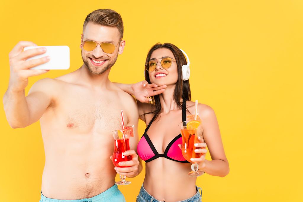hombre feliz en gafas de sol tomando selfie con chica en traje de baño escuchando música y sosteniendo cóctel aislado en amarillo - Foto, Imagen