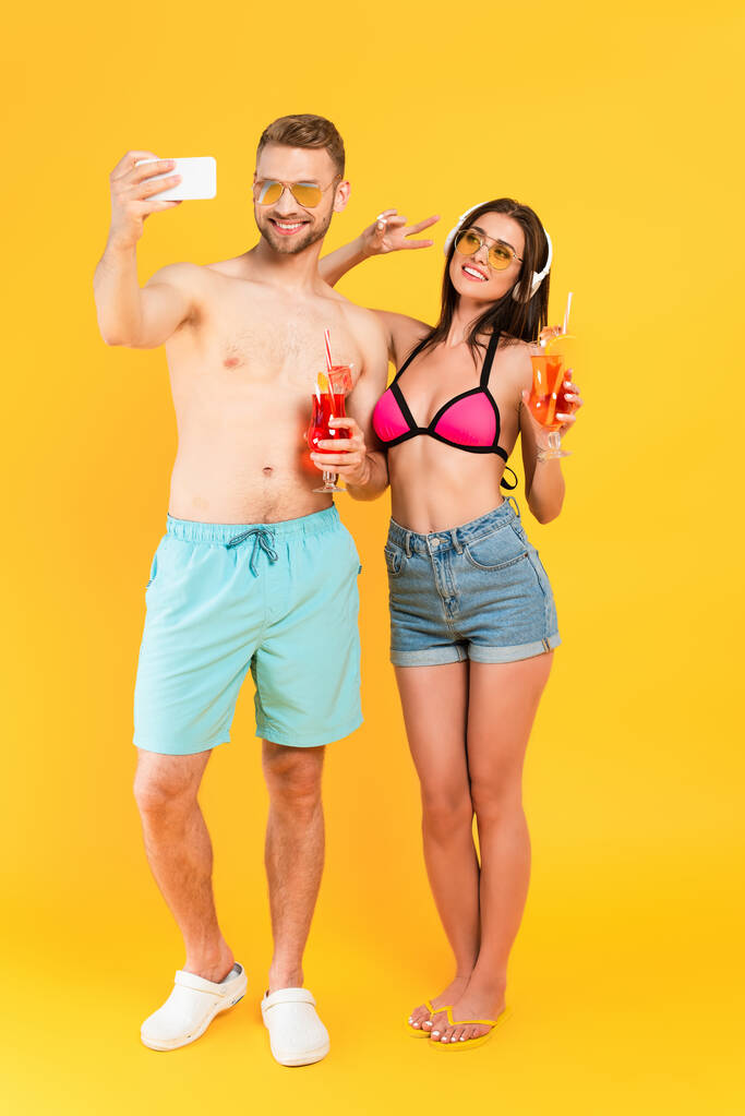 fille heureuse dans les écouteurs montrant signe de paix et de prendre selfie avec l'homme tenant cocktail sur jaune - Photo, image