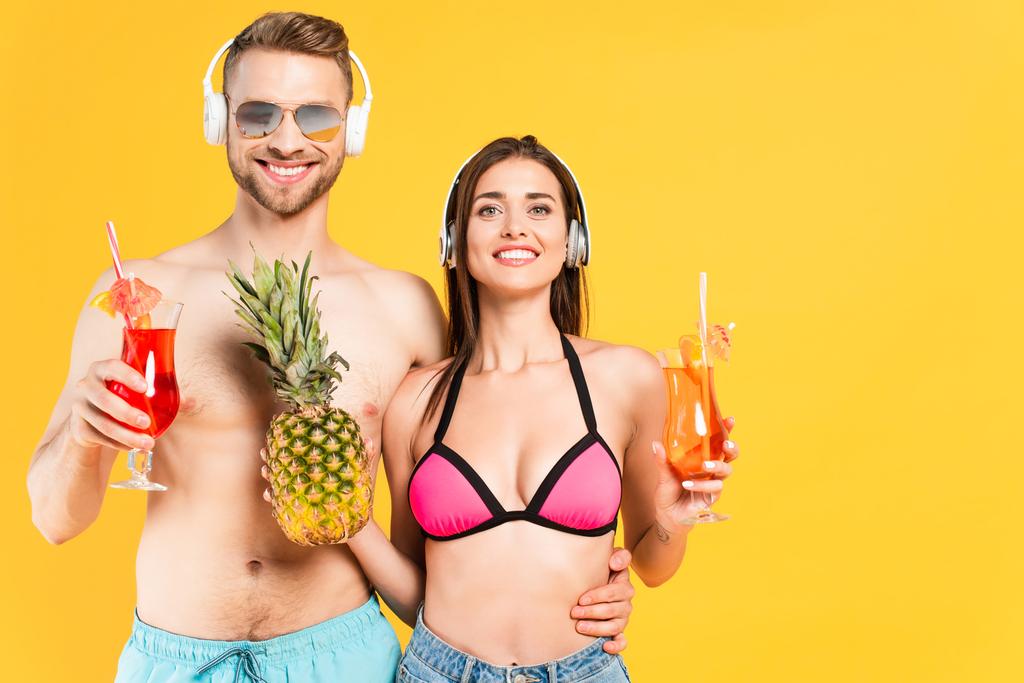 happy man and woman in headphones holding pineapple and cocktails isolated on yellow  - Photo, Image