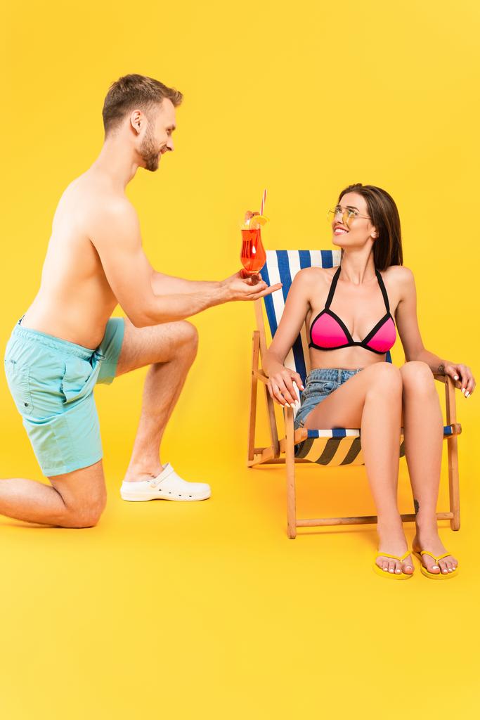 homme torse nu debout sur le genou et tenant cocktail près de fille attrayante et heureuse dans des lunettes de soleil sur jaune - Photo, image
