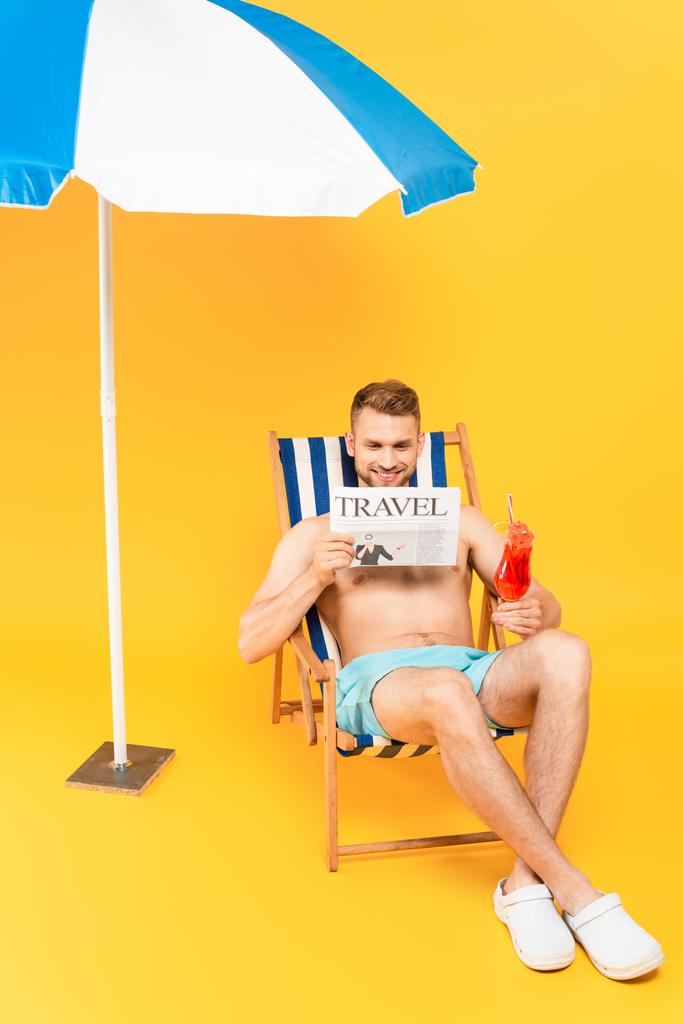 όμορφος άνδρας κάθεται στο κατάστρωμα καρέκλα με κοκτέιλ, ενώ διαβάζετε ταξιδιωτική εφημερίδα κοντά ομπρέλα παραλία στο κίτρινο - Φωτογραφία, εικόνα
