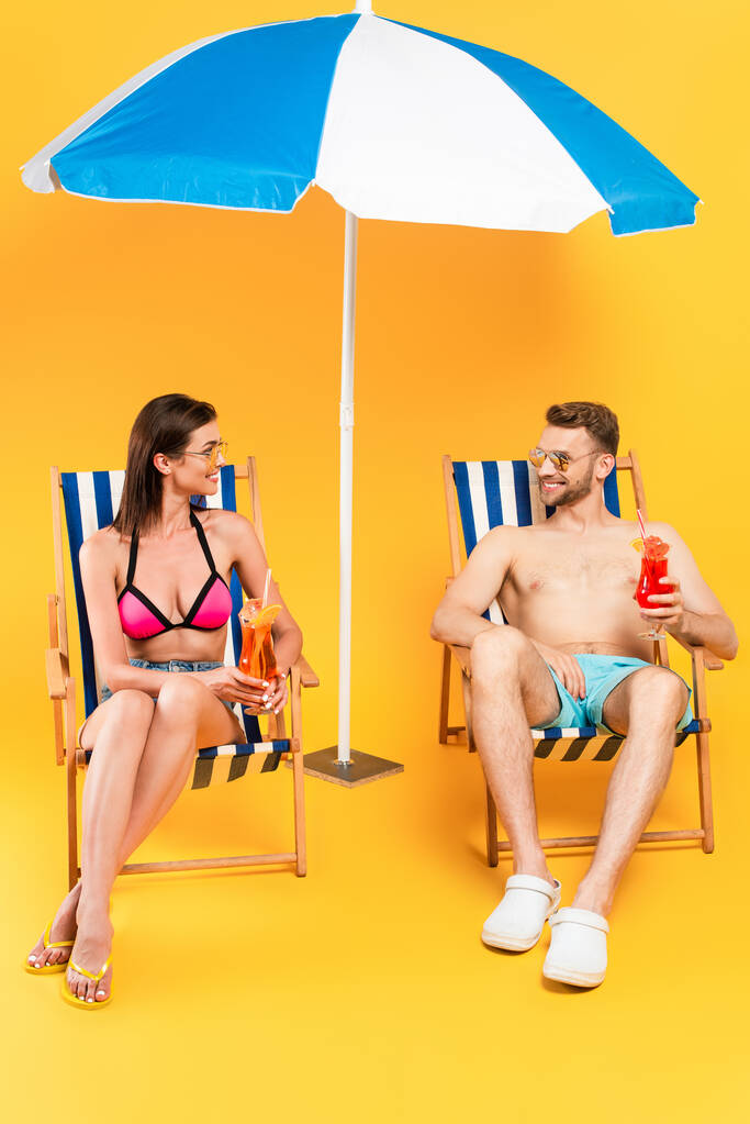 wesoła para w okularach przeciwsłonecznych trzymająca koktajle siedząc na leżakach przy plaży parasol na żółtym - Zdjęcie, obraz