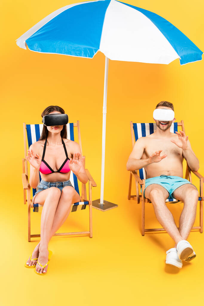 счастливая пара в наушниках виртуальной реальности сидит на шезлонгах и жестикулирует возле пляжного зонта на желтом - Фото, изображение