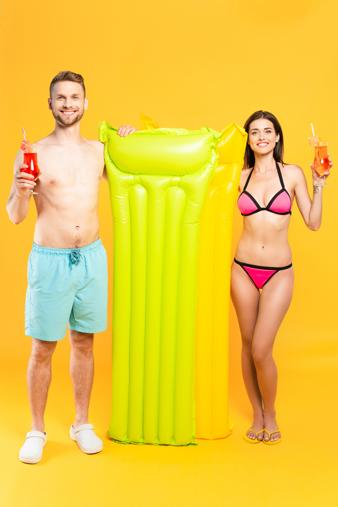 szczęśliwa para w strojach kąpielowych patrząc w kamerę i trzymając koktajle w pobliżu dmuchanego materaca na żółto - Zdjęcie, obraz