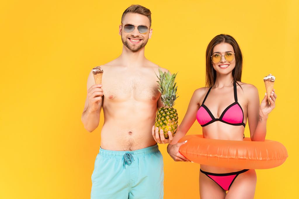 homme musclé heureux avec ananas près de la femme dans des lunettes de soleil tenant la crème glacée tout en se tenant debout avec anneau gonflable isolé sur jaune  - Photo, image