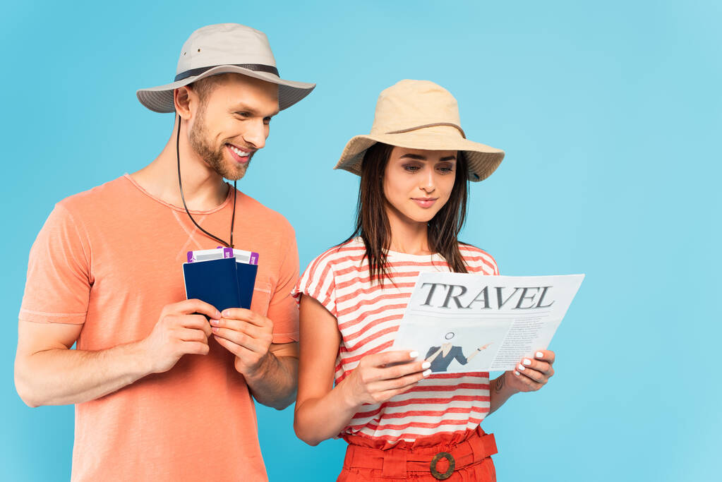 щасливий чоловік у капелюсі тримає паспорти з авіаквитками біля дівчини, читає туристичну газету ізольовано на синьому
 - Фото, зображення
