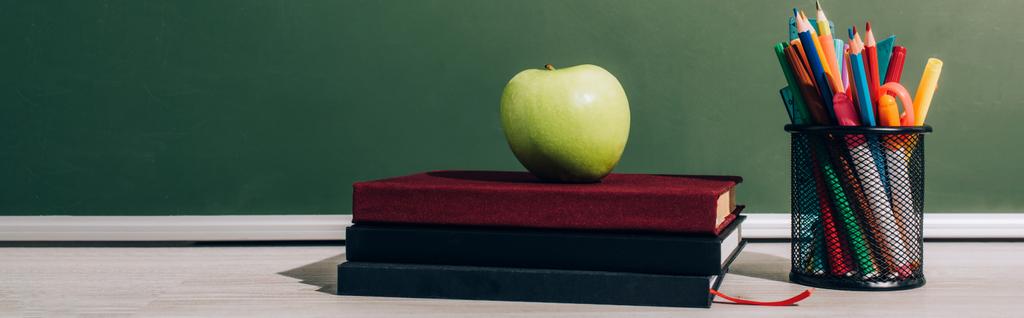 горизонтальное изображение спелого яблока на книгах возле держателя ручки с цветными карандашами возле зеленой доски - Фото, изображение