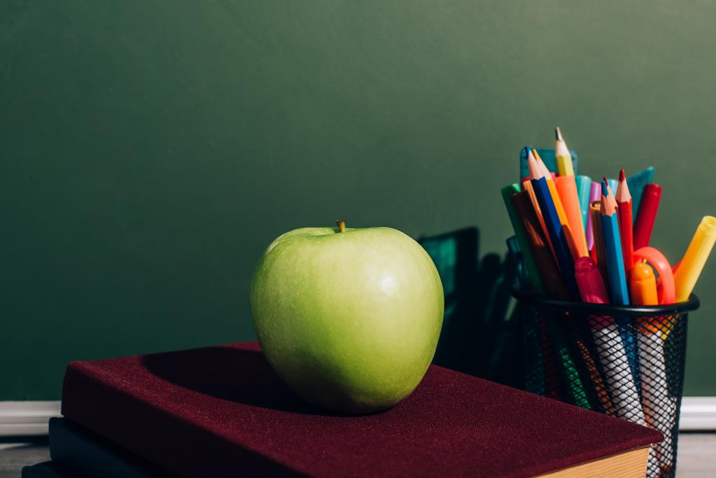 целое яблоко на книгах возле держателя ручки с цветными карандашами и войлочными ручками на столе рядом с зеленой доской - Фото, изображение