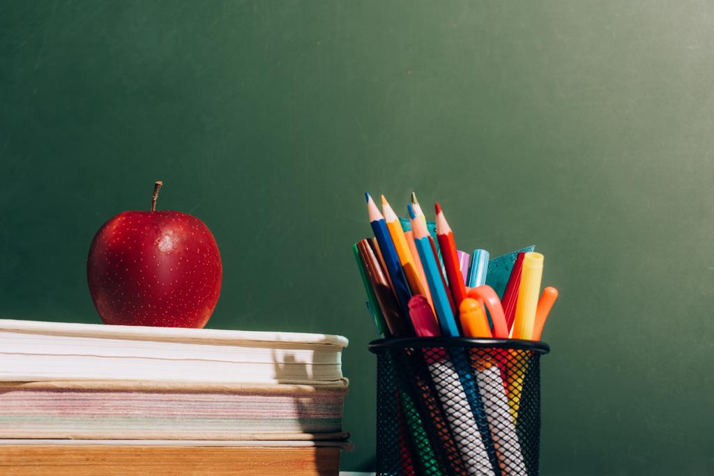 porta-caneta com lápis de cor e canetas de feltro e maçã madura em livros perto de quadro-negro verde
 - Foto, Imagem