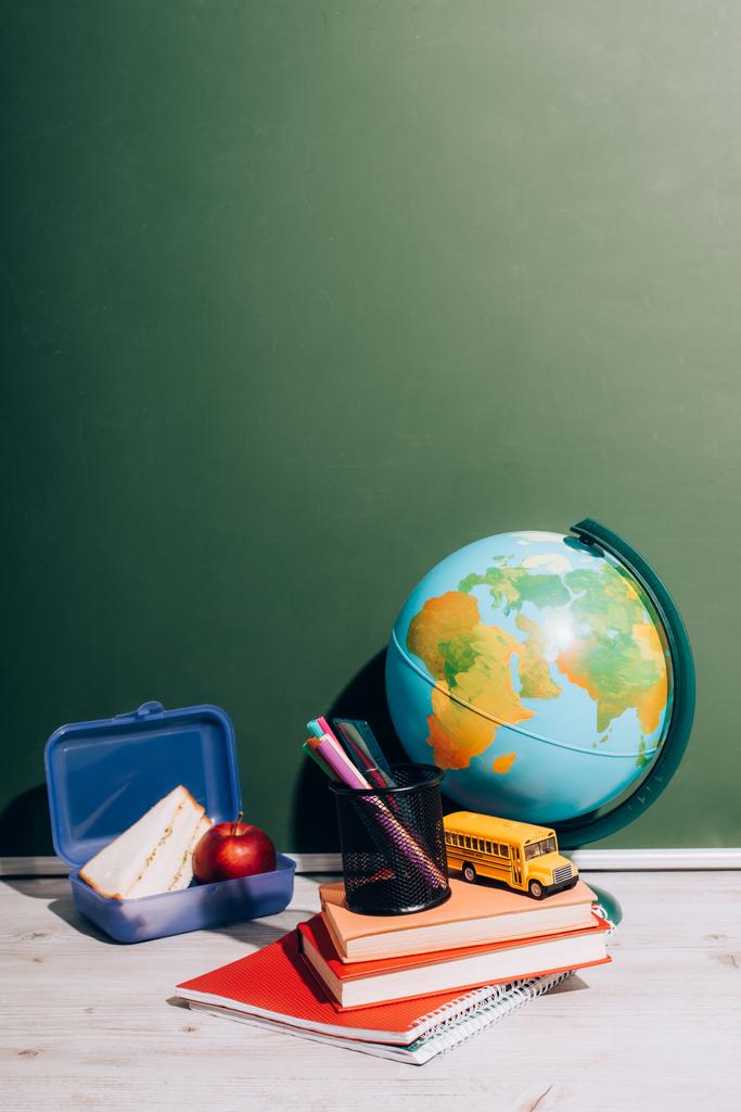 zeměkoule v blízkosti školní autobus model a držák pera na knihy v blízkosti oběda box na stole v blízkosti zelené tabuli - Fotografie, Obrázek