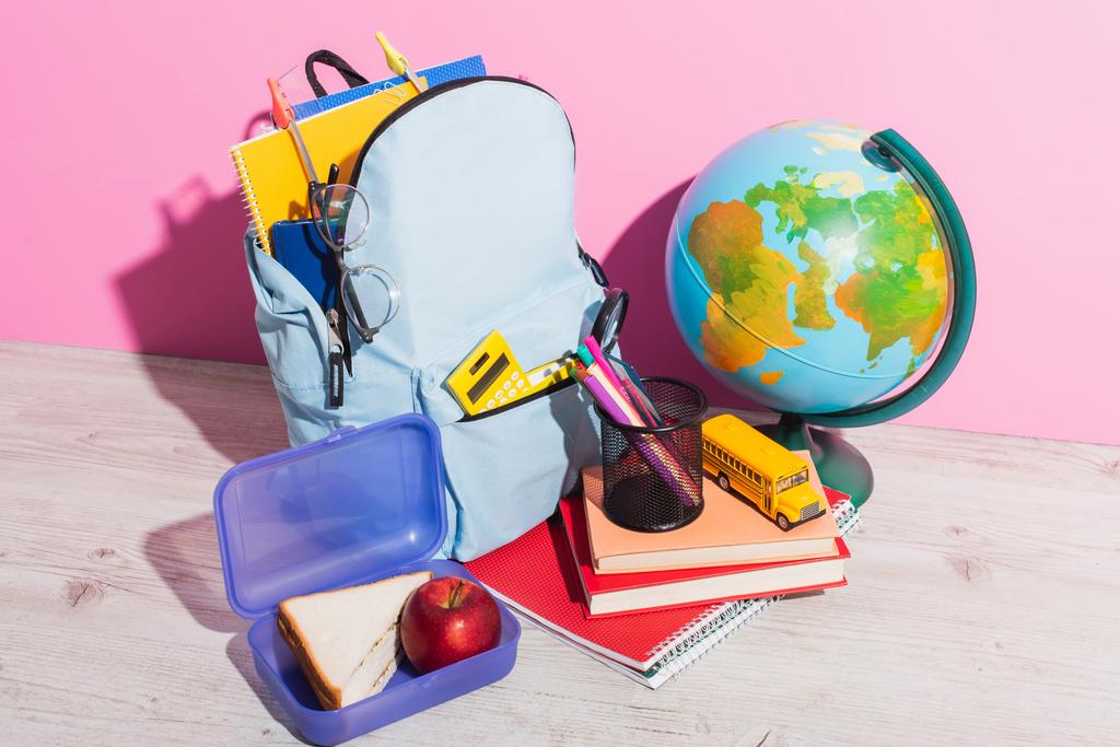 modrý batoh se školními potřebami v blízkosti zeměkoule, občerstvení, knihy, držák pera a školní autobus model na růžové - Fotografie, Obrázek