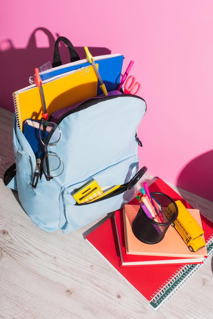 vysoký úhel pohledu na batoh plný školní potřeby v blízkosti knih, držák pera a školní autobus model na růžové - Fotografie, Obrázek