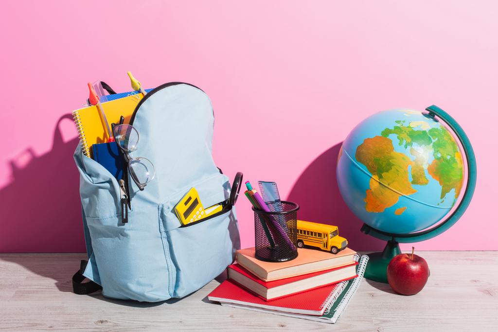 blauer Rucksack mit Schulbedarf in Erdnähe, Büchern, Stiftehalter, frischem Apfel und Schulbusmodell auf rosa - Foto, Bild