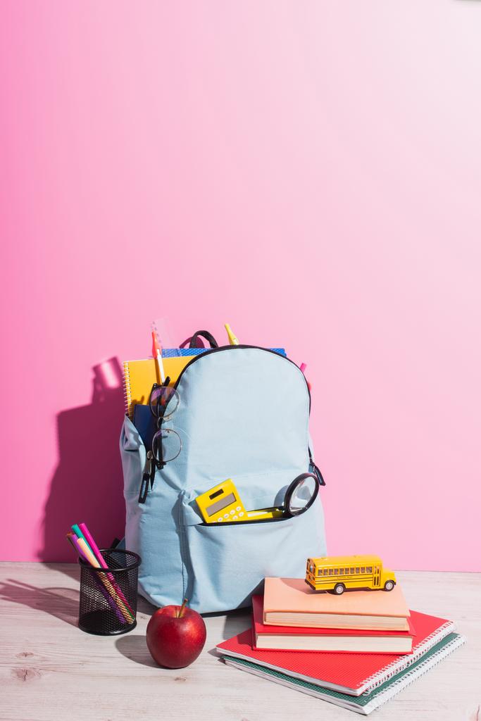 school backpack full of stationery near pen holder, ripe apple, books and school bus model on pink - Photo, Image