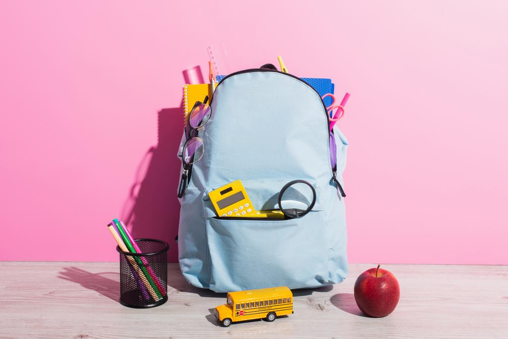 sac à dos bleu emballé avec papeterie scolaire près du modèle d'autobus scolaire, livres, pomme mûre et porte-stylo sur rose - Photo, image