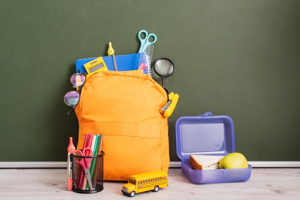 Желтый рюкзак со школьными принадлежностями рядом с коробкой для обеда, школьной моделью автобуса и держателем ручки на столе рядом с зеленой доской - Фото, изображение