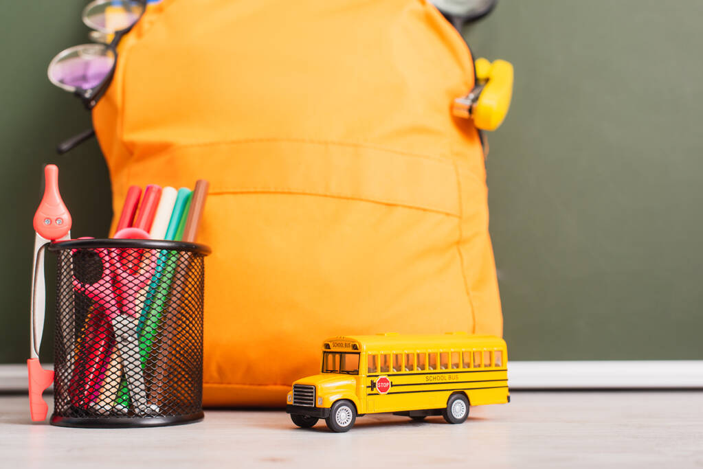 Желтый рюкзак рядом с моделью школьного автобуса, держатель ручки с войлочными ручками, ножницами и разделителем компаса на столе возле зеленой доски - Фото, изображение