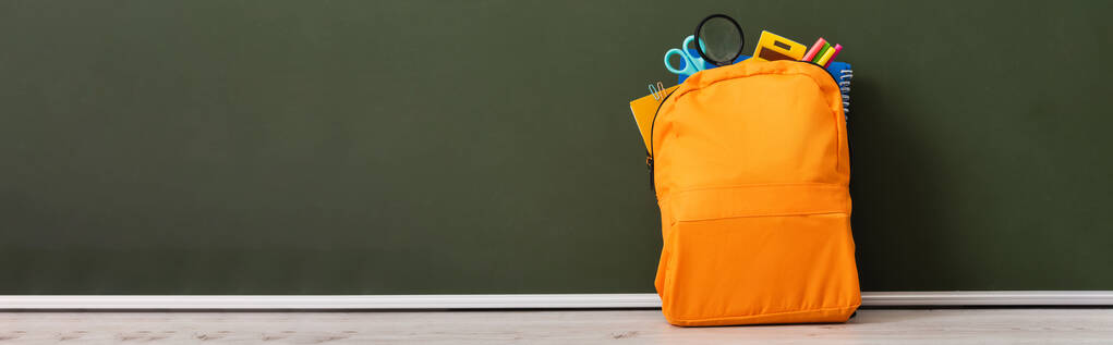 imagen horizontal de la mochila amarilla llena de papelería escolar en el escritorio cerca de pizarra verde
 - Foto, Imagen