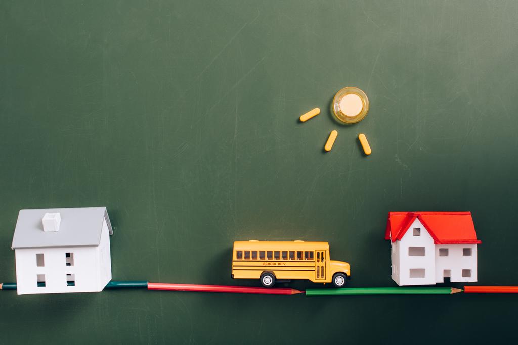 верхний вид моделей домов, игрушечный школьный автобус на дороге, сделанный из цветных карандашей, и солнце из магнитов на зеленой доске - Фото, изображение