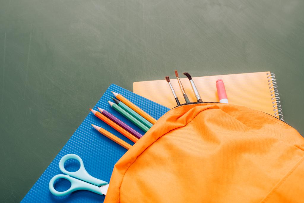 Yeşil karatahtanın üzerinde okul malzemeleri olan sarı sırt çantasının yüksek açılı görüntüsü - Fotoğraf, Görsel