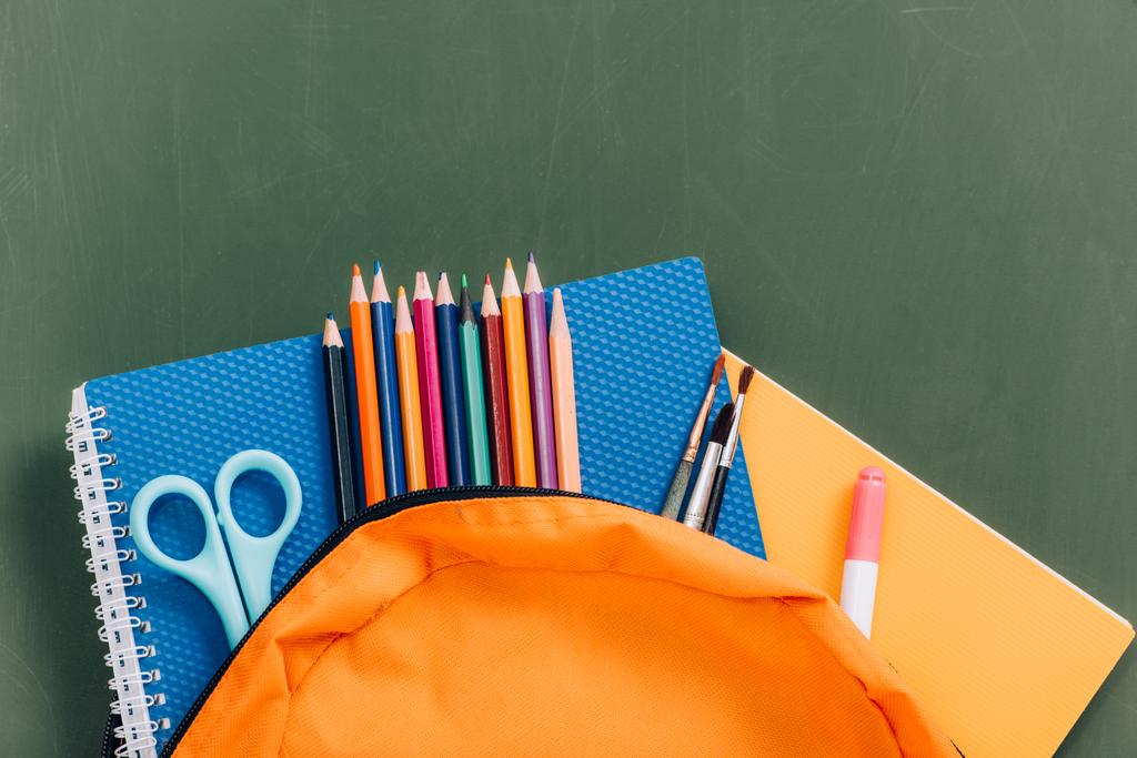 bovenaanzicht van gele rugzak met kleurpotloden, notitieboekjes, schaar en penselen bij groen schoolbord - Foto, afbeelding