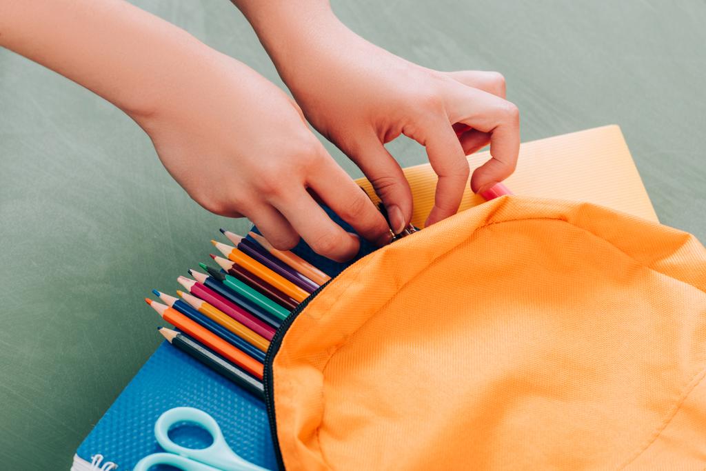 przycięty widok ucznia biorącego papeterię z plecaka z notatnikami, kolorowymi ołówkami i nożyczkami na zielonej powierzchni - Zdjęcie, obraz