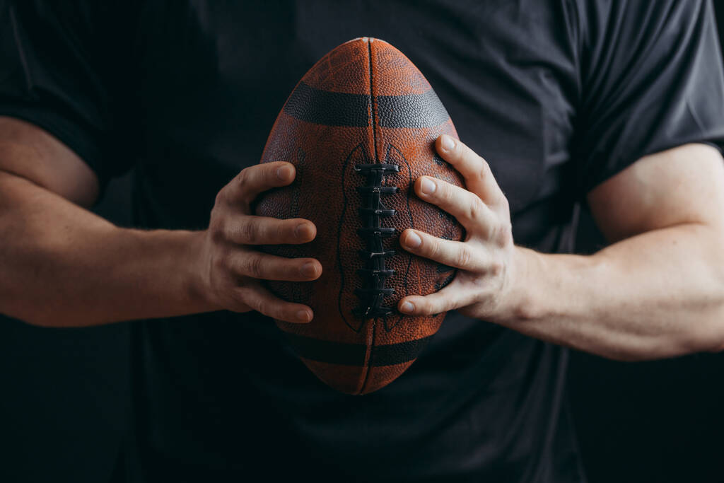 Jogador de futebol americano muscular em uniforme protetor e capacete segurando bola
 - Foto, Imagem