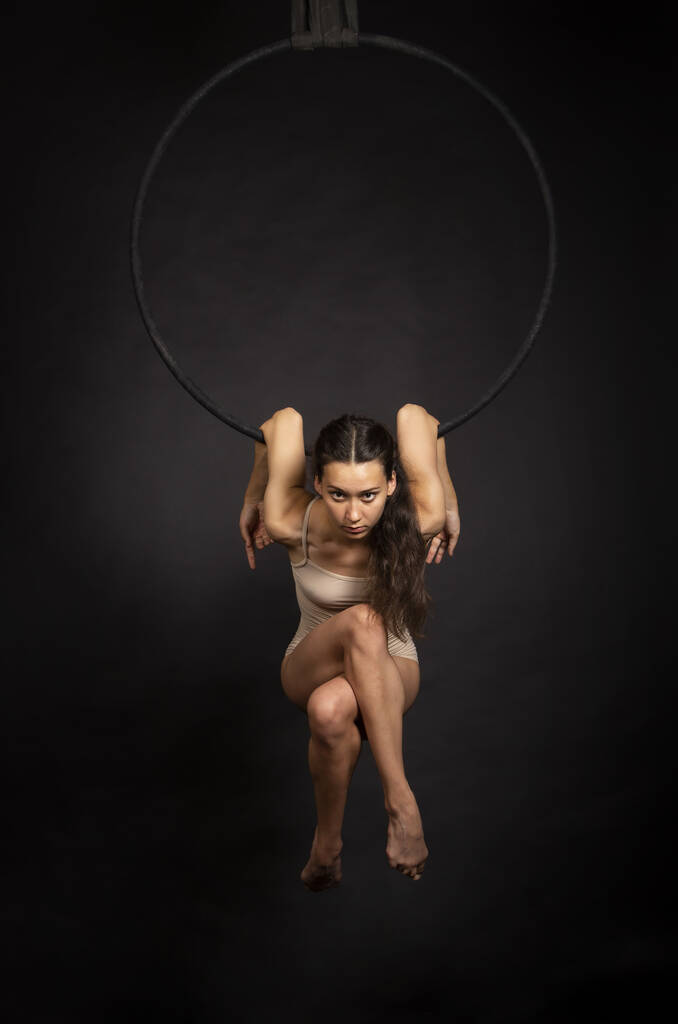Une jeune femme aux cheveux longs est engagée dans des acrobaties aériennes en body beige, effectue des exercices dans l'anneau aérien. Studio tournage sur un fond sombre. - Photo, image