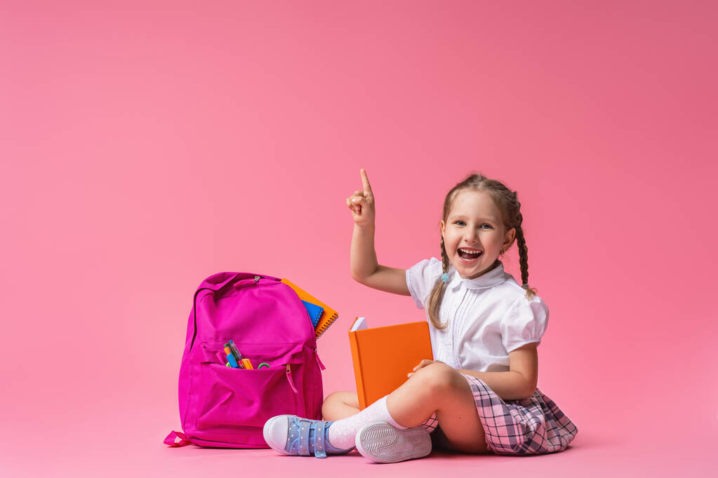 szczęśliwa uśmiechnięta dziewczynka w mundurze z plecakiem i książkami siedzi na różowym tle w Studio. Dziecko wskazuje palcem w górę. Wracać do szkoły. przestrzeń kopiowania - Zdjęcie, obraz