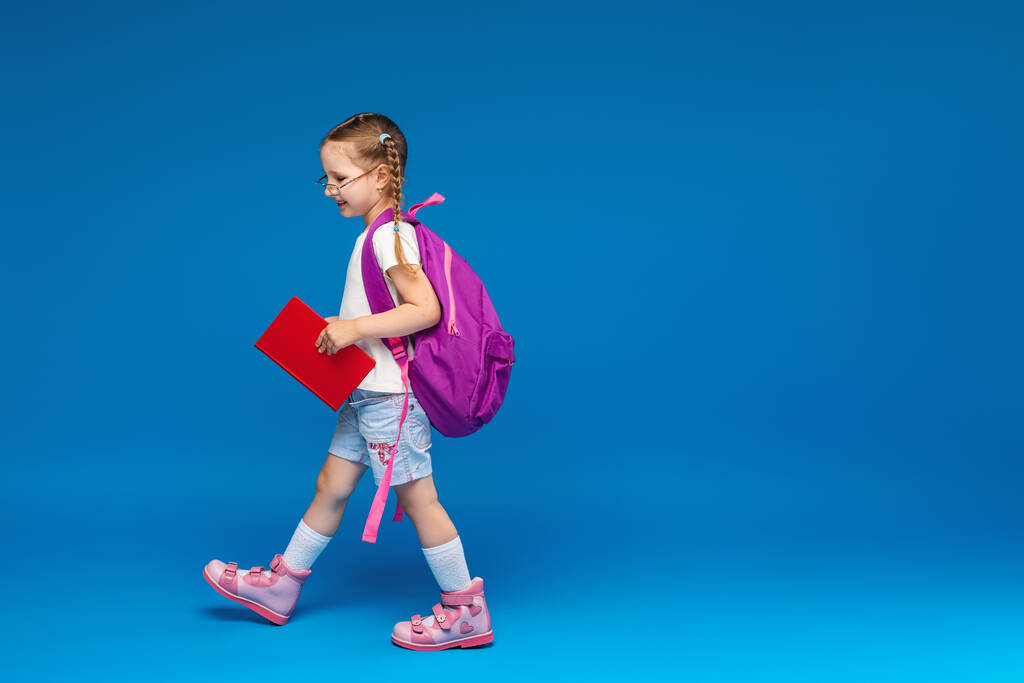 Joyeux sourire petite fille va à l'école pour la première fois. Un enfant avec un cartable et un livre à la main sur un fond bleu. Enfant dans la classe. Retour à l'école. - Photo, image