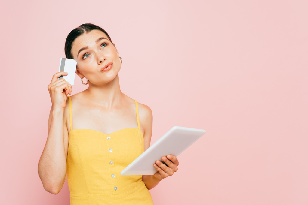 zamyšlená brunetka mladá žena s digitálním tabletem a kreditní kartou izolované na růžové - Fotografie, Obrázek