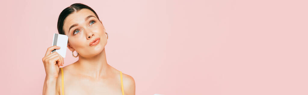 задумчивая брюнетка молодая женщина с кредитной картой изолированы на розовый, панорамный снимок - Фото, изображение