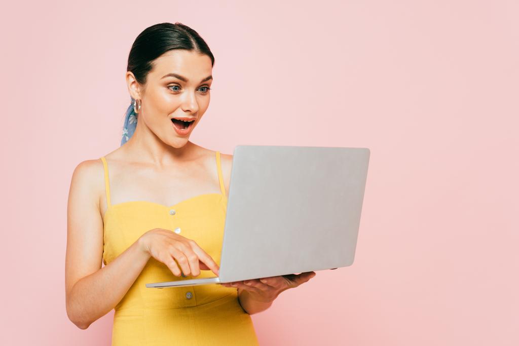 sorpreso bruna giovane donna utilizzando computer portatile isolato su rosa - Foto, immagini