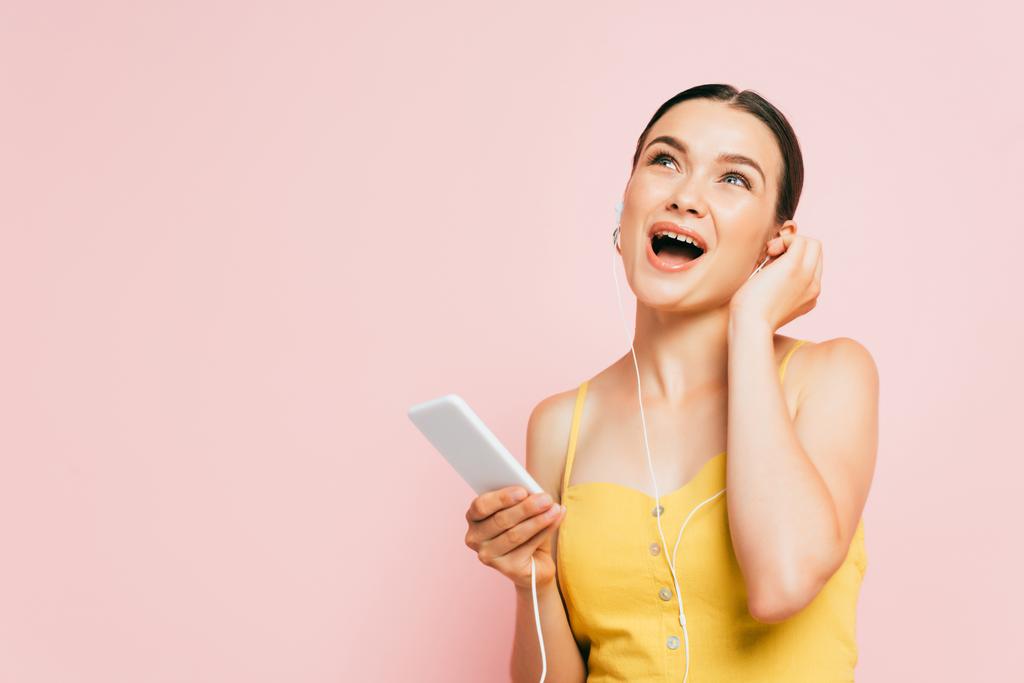 興奮したブルネットの若い女性がイヤフォンで音楽を聞いてピンクに隔離されたスマートフォンを保持 - 写真・画像