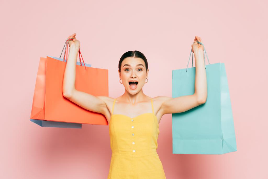 excited brunette young woman with shopping bags in raised hands on pink - Photo, Image